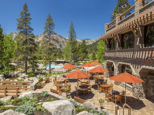 An outdoor dining area with wooden tables and orange umbrellas, set in a lush, green mountain landscape next to a building sign reading 'Sandy's Pub'.