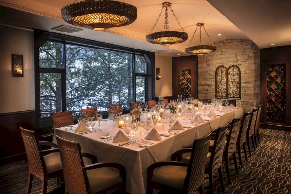 A beautifully set dining table in an elegant room, featuring folded napkins, wine glasses, and lit candles, with large windows offering an outdoor view.