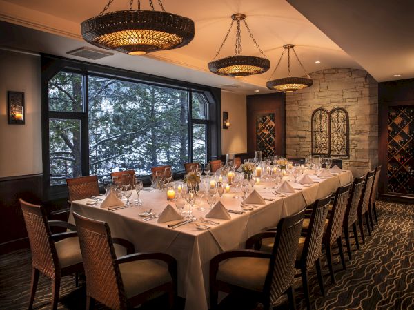 A beautifully set dining table in an elegant room, featuring folded napkins, wine glasses, and lit candles, with large windows offering an outdoor view.