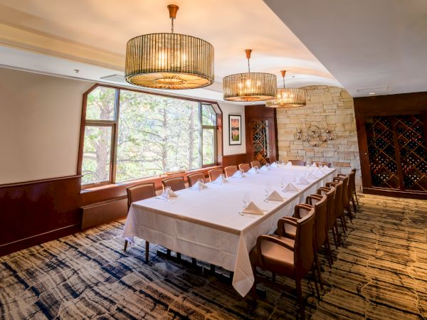 A restaurant dining room set with a long table, upholstered chairs, hanging chandeliers, and a stone fireplace wall.