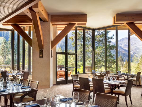 A modern lodge dining room with set tables, large windows, and vaulted wooden beams overlooking pine-covered mountains.