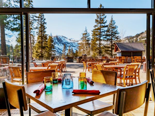 The image shows a dining area in a restaurant with wooden tables and chairs, set against a stunning mountain and forest backdrop with snow.