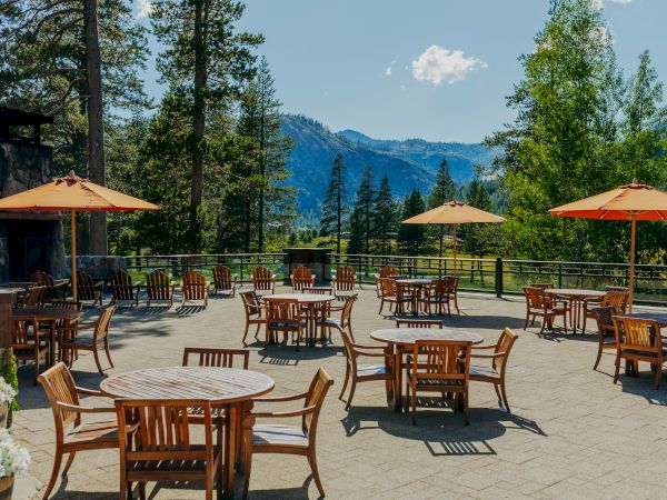 An outdoor patio with tables, chairs, and large umbrellas overlooks a scenic mountain view surrounded by tall trees and a clear sky.