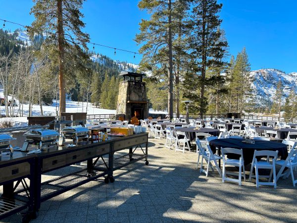Outdoor dining area with tables and chairs set up, buffet station, surrounded by trees and snowy mountains. Bright, sunny day.