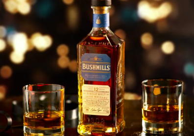 A bottle of Bushmills Irish whiskey flanked by two glasses of whiskey on a bar, warm bokeh lights in the background.