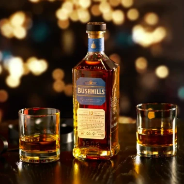 A bottle of Bushmills Irish whiskey flanked by two glasses of whiskey on a bar, warm bokeh lights in the background.