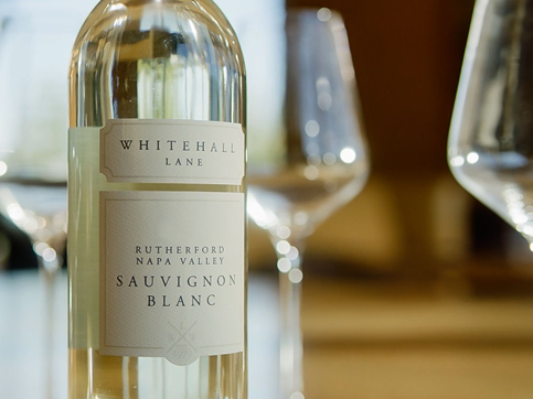 A bottle of Whitehall Lane Sauvignon Blanc with two filled wine glasses in the background, on a wooden table.