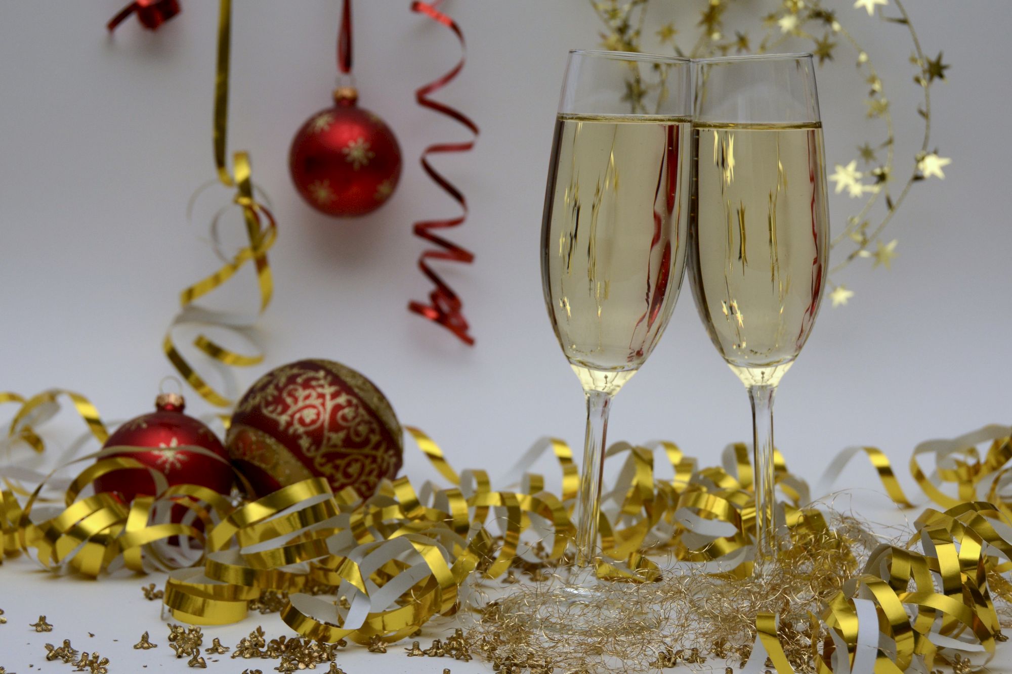 Two champagne glasses, red and gold Christmas ornaments, and gold streamers on a white background, creating a festive celebration scene.