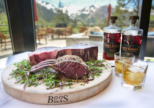 A cutting board with raw steaks and seasonings, two bottles of Frey Ranch bourbon, and two glasses of whiskey, with a scenic mountain view.