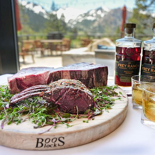 A cutting board with raw steaks and seasonings, two bottles of Frey Ranch bourbon, and two glasses of whiskey, with a scenic mountain view.