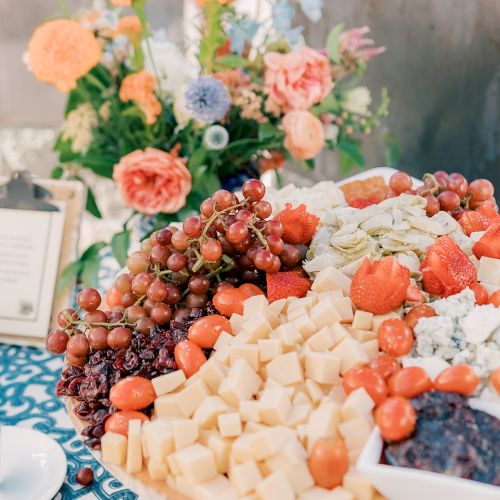 A platter with assorted cheeses, grapes, and cherry tomatoes, set on a table with a floral arrangement and a clipboard.