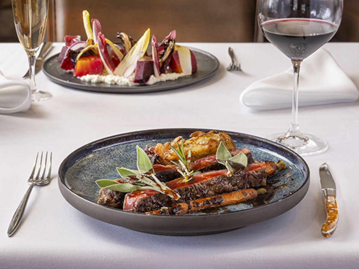 A dish with steak and vegetables is in the foreground, with a beet salad and wine glasses on a white tablecloth in the background.