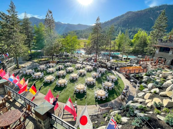 The image shows an outdoor event setup with multiple round tables and international flags, set amidst a scenic mountain landscape with evergreen trees and rocks.