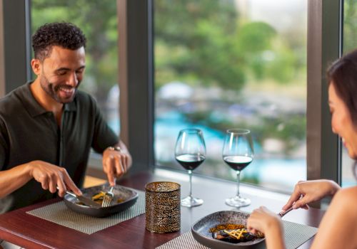 Two people are dining together at a table with two glasses of red wine, enjoying their meal in a cozy setting, near large windows.