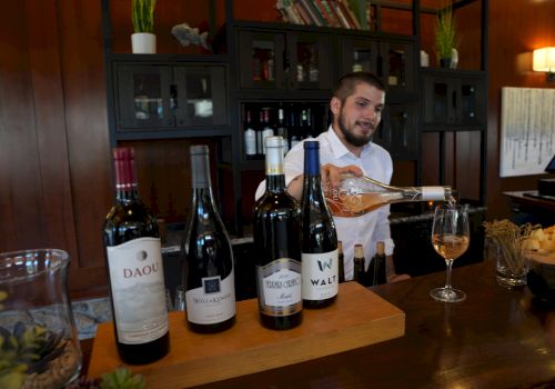 A chef pours wine at a tasting counter with several bottles lined up on a wooden board in a cozy, wood-paneled room.