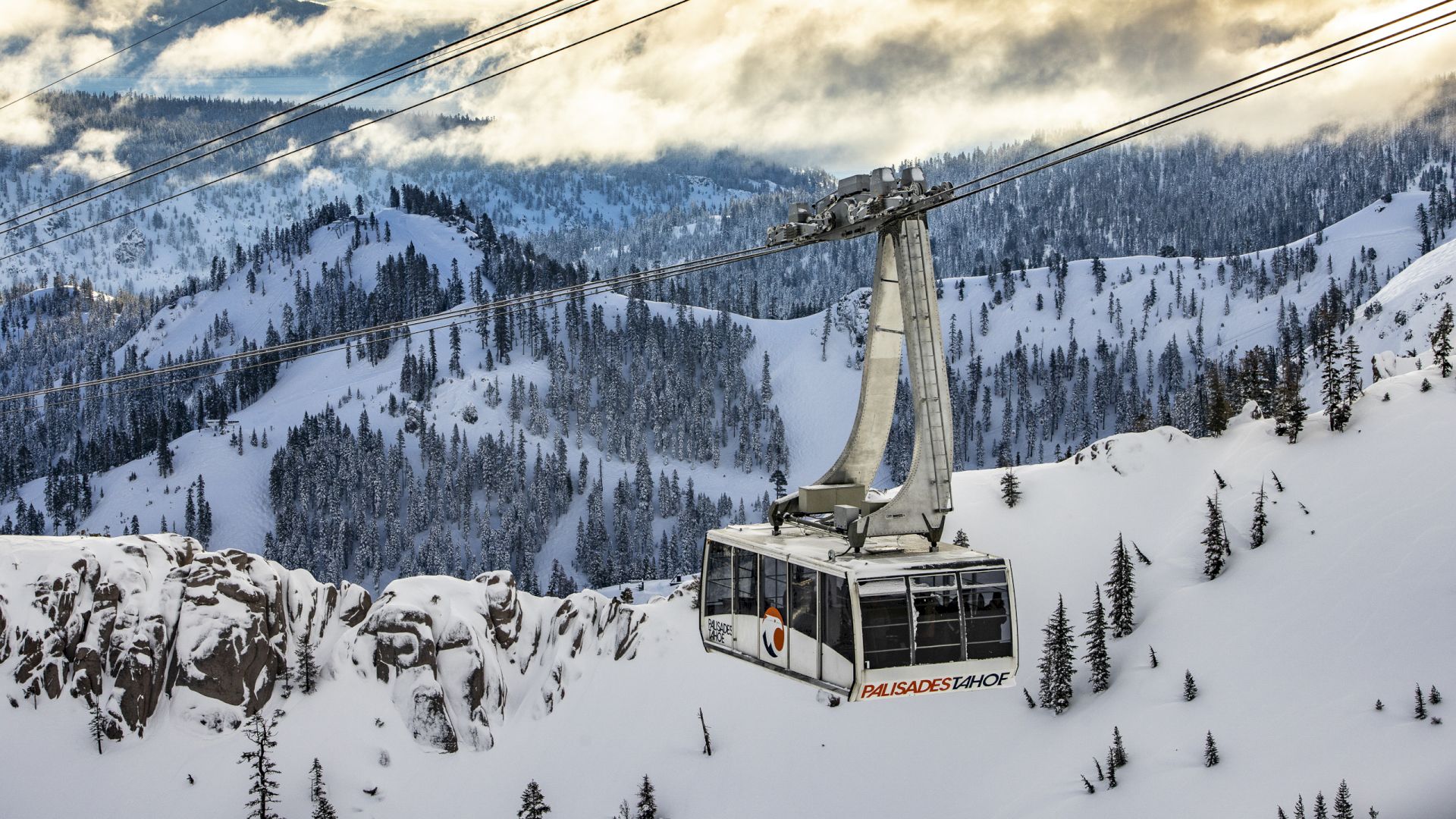 A cable car travels over a snowy mountain landscape under a partly cloudy sky, surrounded by forested slopes.