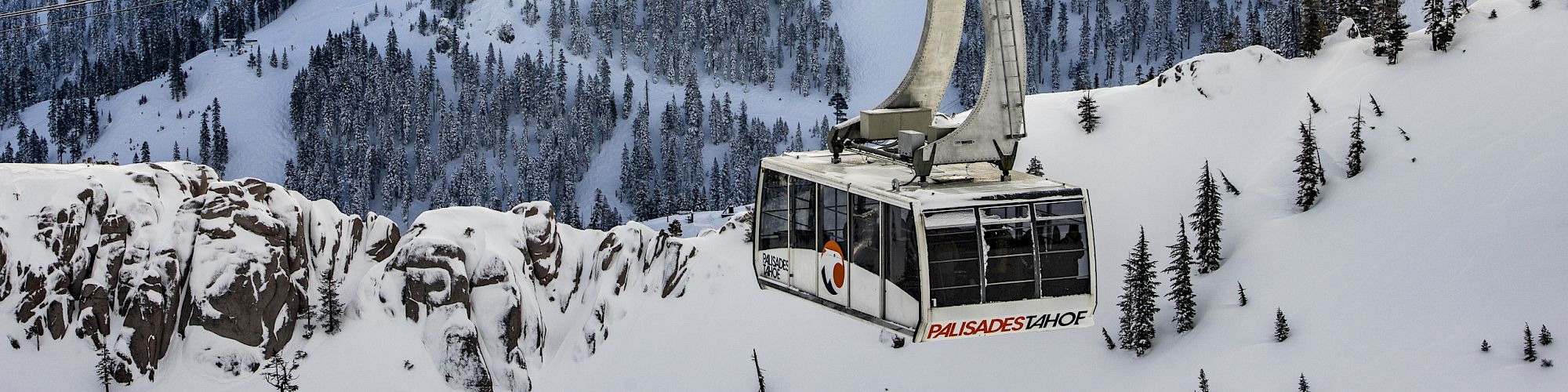 A cable car travels over a snowy mountain landscape under a partly cloudy sky, surrounded by forested slopes.