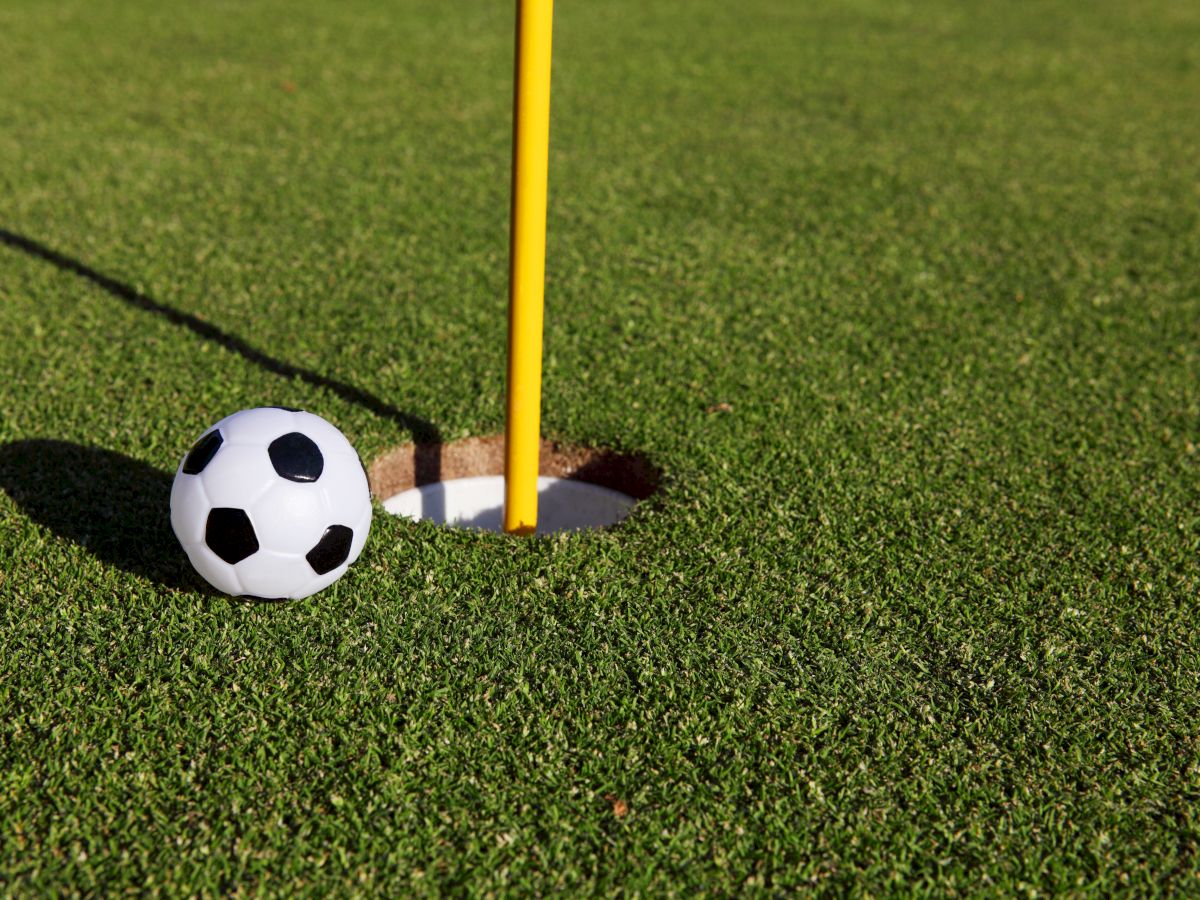 A soccer ball is positioned next to a hole on a golf course, with a flagpole in the hole, blending elements of both sports perfectly.