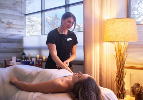 A spa or massage therapist works on a client lying face up on a table in a cozy room with warm lighting and large windows ending with a soft, relaxed atmosphere.