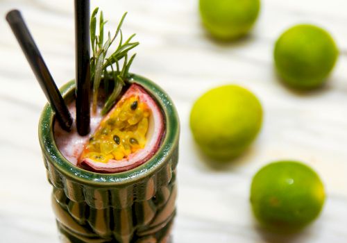 A tropical-looking drink served in a green cup with a watermelon slice and rosemary, two black straws, plus limes in the background.