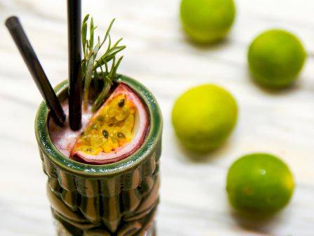 A tiki mug with a watermelon slice straw, rosemary sprig garnish, and lime halves in the background, plus a watermelon wedge nearby.