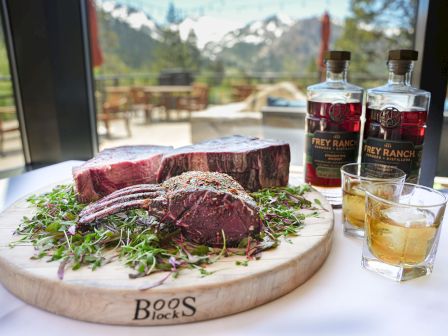 A sliced steak on a wooden board with herbs, two bottles of whiskey beside, and a glass of amber liquor on a sunny outdoor table.