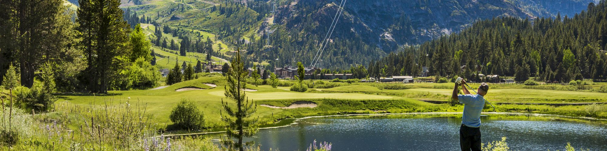 A person is playing golf in a scenic mountainous area, with lush greenery, a pond, and clear blue sky in the background.