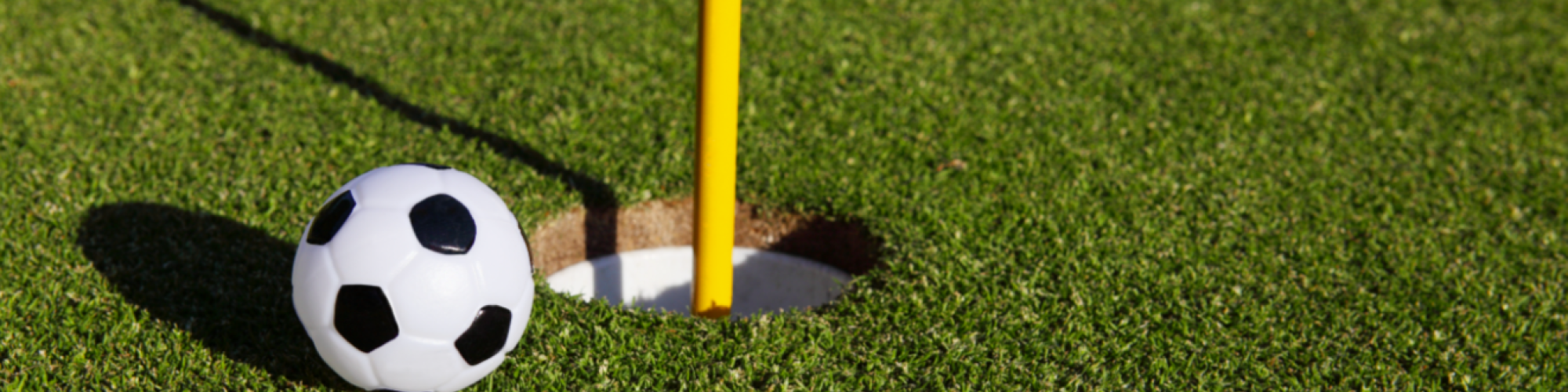 The image shows a small soccer ball next to a hole on a golf course, with a yellow flagstick in the hole.