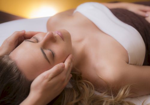 A person is lying down with eyes closed, receiving a relaxing facial massage from a pair of hands while wrapped in a white towel.