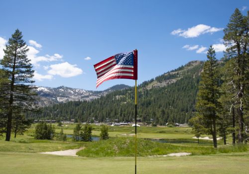 An American flag is flying on a golf course with a backdrop of lush green mountains and trees under a clear blue sky with some white clouds.