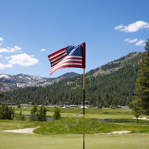 An American flag is flying on a golf course with a backdrop of lush green mountains and trees under a clear blue sky with some white clouds.