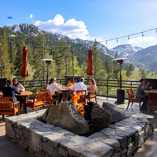 People sitting around a fire pit on an outdoor patio with mountains and trees in the background, under a clear sky with fairy lights strung above.