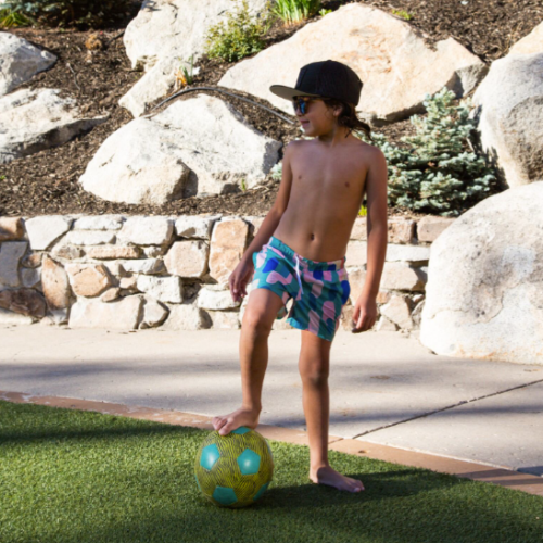 A child wearing shorts, sunglasses, and a hat stands on grass with one foot on a soccer ball, with rocks and plants in the background.