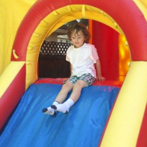 A child with curly hair, wearing a white shirt and checked shorts, is sliding down an inflatable slide in a colorful play area.