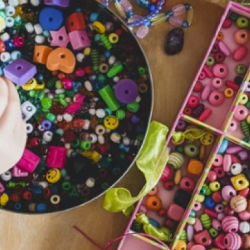 A hand is selecting beads from a bowl filled with colorful beads, with a pink organizer containing more sorted beads and materials on the side.