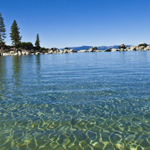 The image shows a serene lake with clear water, rocky shoreline, and trees in the background under a clear blue sky.