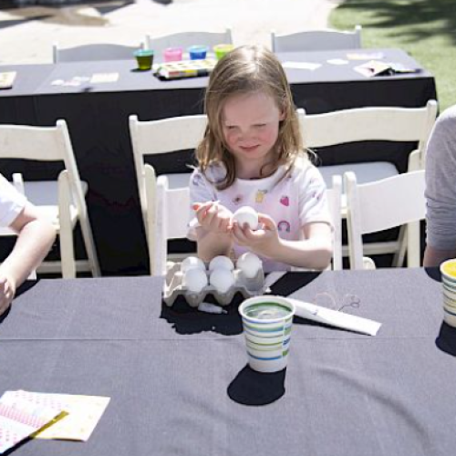 Children are sitting at a table with art supplies, eggs, and paint cups, appearing to be engaged in a craft activity outdoors.