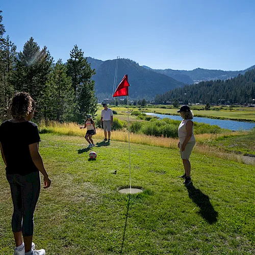 A group plays golf on a grassy course beside a pond, with a red flag, trees, and mountains in the sunny background.
