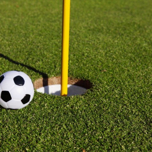 A small soccer ball is next to a hole on a golf course, with a yellow flagstick inserted in the hole. The scene is on a lush green putting green.