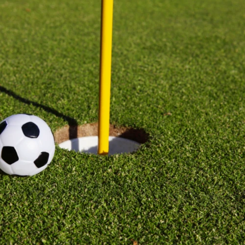 A small soccer ball is next to a hole on a golf course, with a yellow flagstick inserted in the hole. The scene is on a lush green putting green.