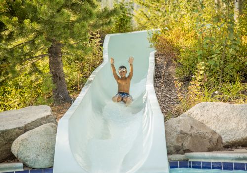 A child wearing swim goggles is sliding down a water slide with arms raised, surrounded by greenery and rocks, heading toward the pool below.