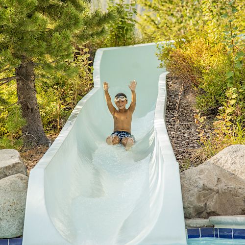 A child wearing swim goggles is sliding down a water slide with arms raised, surrounded by greenery and rocks, heading toward the pool below.