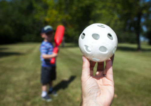 A person is holding a plastic ball with holes, in front of a child holding a red plastic bat, standing on a grassy field, ready to play baseball.