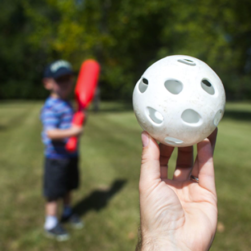 A person is holding a plastic ball with holes, in front of a child holding a red plastic bat, standing on a grassy field, ready to play baseball.