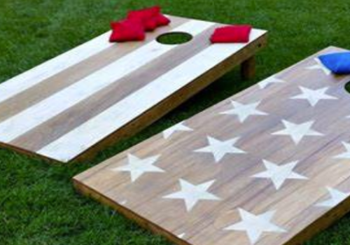 The image shows two cornhole boards with American flag designs, placed on grass, accompanied by red and blue bean bags.