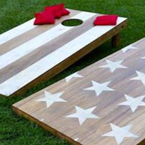 The image shows two cornhole boards with American flag designs, placed on grass, accompanied by red and blue bean bags.