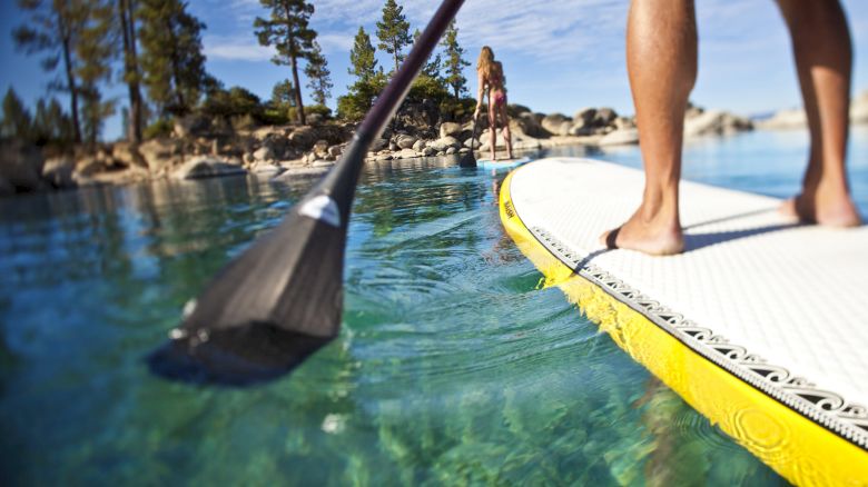 A person is paddleboarding on clear, turquoise water with trees and rocks in the background. Another person stands onshore in the distance.