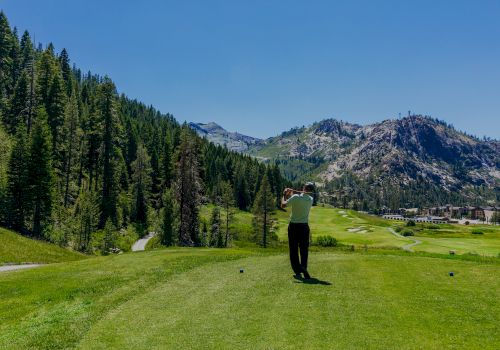 A person is playing golf on a lush course surrounded by mountains and trees under a clear blue sky.