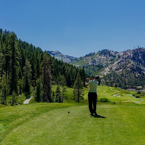 A person is playing golf on a lush course surrounded by mountains and trees under a clear blue sky.