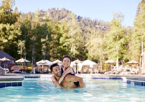 A couple enjoys time together in a pool, with scenic trees and mountains in the background on a sunny day, surrounded by lounge chairs and umbrellas.